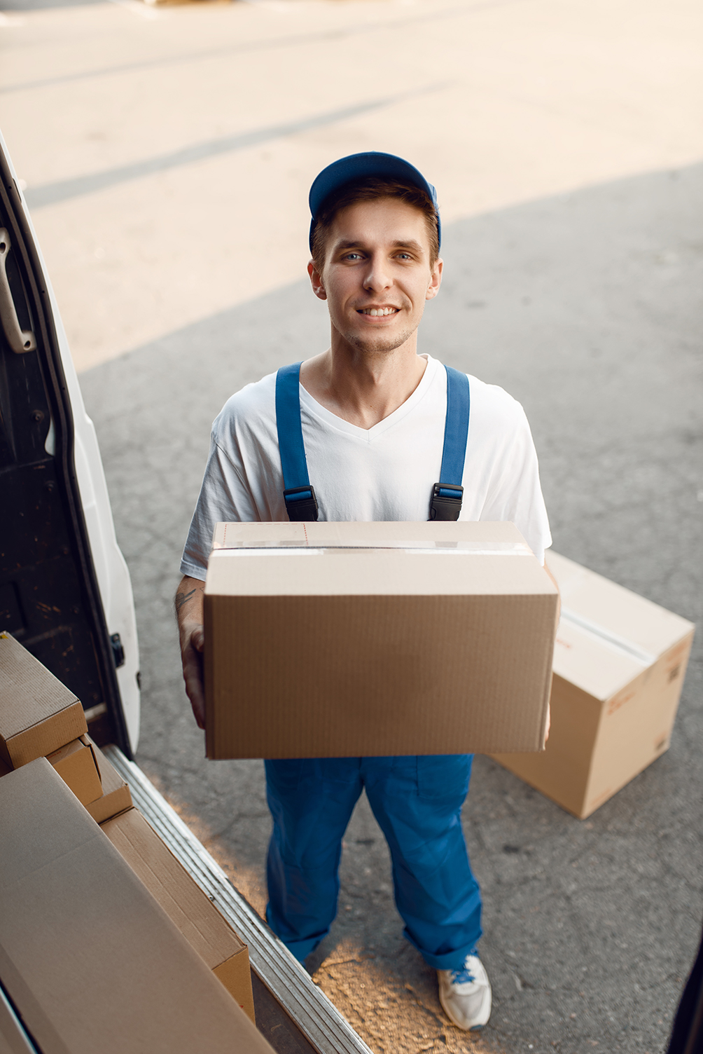 smiling-delivery-worker-in-blue-overalls-holding-box-near-van
