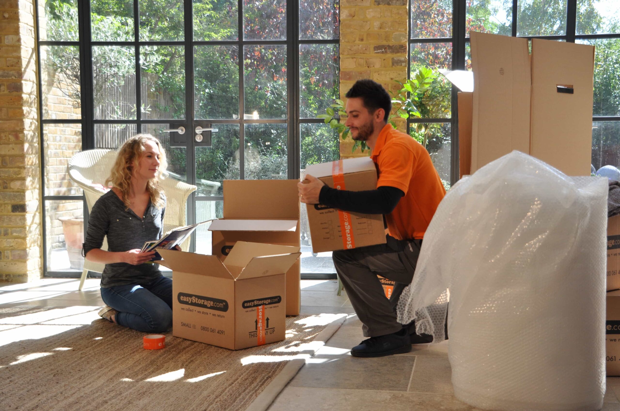 Mover-in-orange-shirt-holding-box-in-living-room,-with-another-person-sitting-next-to-open-box,-large-windows-in-background.