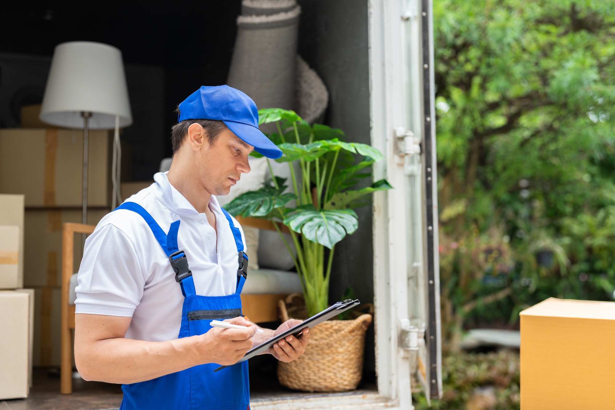 Man mover in blue uniform checking lists on clipboard while unloading cardboard boxes from truck