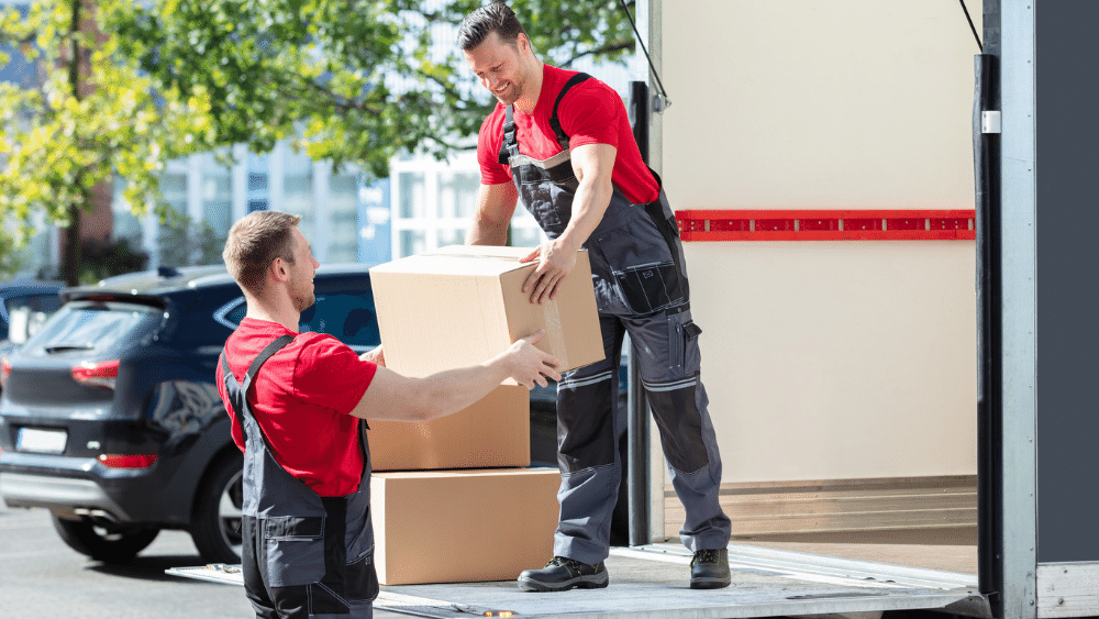 Two-delivery-workers-wearing-red-t-shirts-and-overalls,-loading-cardboard-boxes-onto-a-delivery-truck-in-sunny-outdoor-environment.