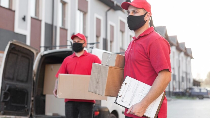 Two-delivery-workers-wearing-red-t-shirts,-caps,-and-face-masks,-holding-boxes-and-documents-in-front-of-a-delivery-van.