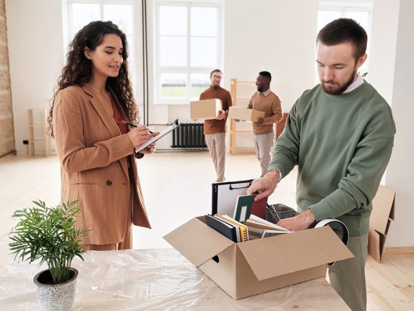 A-woman-holding-a-clipboard-smiling-while-observing-a-person-packing-items-into-a-box-with-two-people-carrying-boxes-in-the-background